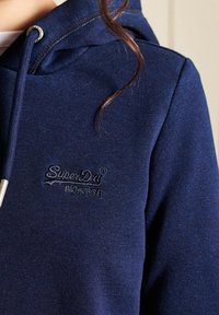 Sudadera azul marino con un acabado texturizado, que presenta un logo bordado de "SuperDry" en el pecho y una capucha con cordones y acentos metálicos.