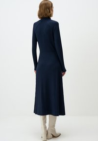 Marineblauwe geribbelde gebreide jurk met een hoge kraag, lange mouwen, een aangesloten taille en een uitlopende zoom, gecombineerd met crèmekleurige kniehoogte haklaarzen.