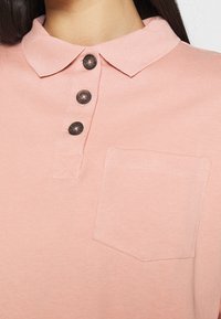 Hellrosa Poloshirt mit kragenartigem Ausschnitt, drei dunklen Knöpfen und einer linken Brusttasche mit einfachen Ziernähten.