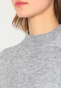 Jasnoszary, dziergany sweter z wysokim, prążkowanym kołnierzem, miękką fakturą i subtelnym melanżowym wzorem. Skupienie na okolicy szyi oraz detalu tkaniny.