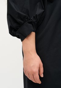 Ανοιχτόχρωμο χέρι χαλαρό στο πλάι, φορώντας μαύρο μακρυμάνικο με ελαστικό μανσέτα, πάνω σε απλό φόντο.