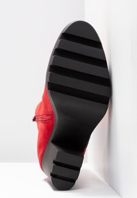 Suela negra con estrías de una bota de tobillo de ante rojo con cremallera lateral, posicionada verticalmente contra un fondo blanco en la esquina.