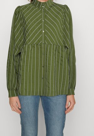 Blusa verde con botones, con rayas blancas verticales y diagonales, cintura y puños fruncidos, llevada con vaqueros azules.