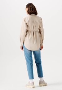 Beige Hemdbluse mit Gürtel, abgerundetem Saum, langen Ärmeln und Knopfmanschetten, kombiniert mit hellblauen Jeans und neutralen Sneakers.