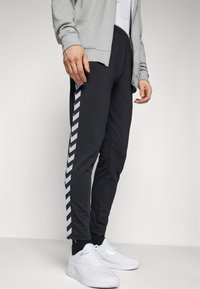 Zwarte joggers met een grijs-witte chevronstreep aan de zijkant, gecombineerd met een lichtgrijze zip-up jas en witte sneakers.