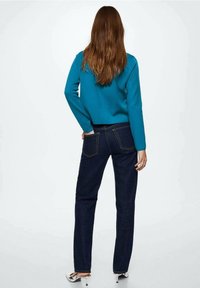 Türkiser Pullover mit langen Ärmeln, kombiniert mit dunkelblauen Jeans mit Gesäßtaschen und goldenen Nähten. Modell von hinten, das die Passform des Outfits zeigt.