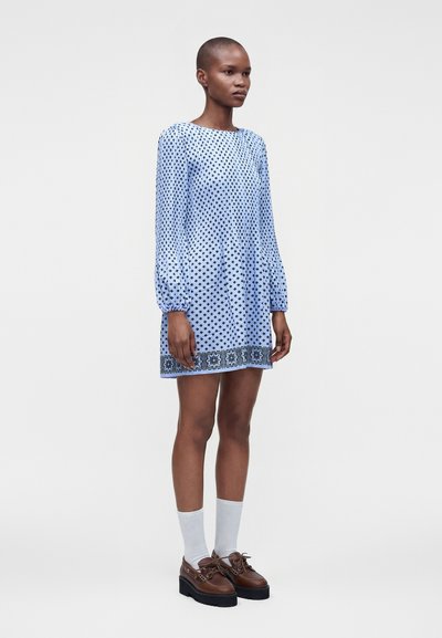 Femme portant une mini-robe bleue à manches longues avec un motif géométrique noir, des chaussettes blanches et des chaussures compensées marron, debout sur un fond uni.