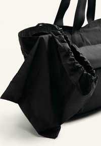 Schwarze Tragetasche aus glattem, leichtem Material mit gerafften Akzenten und stabilen Stoffgriffen. Enthält eine weiche, faltbare Tasche.