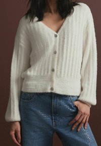 Un cardigan blanc côtelé avec un décolleté en V profond et cinq boutons ornés de pierres précieuses ; il présente un tissu doux et texturé ainsi que de larges manches, associé à un jean bleu.