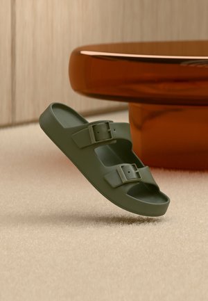 Sandalia tipo chancla verde oliva con dos hebillas ajustables, flotando sobre una alfombra beige junto a una mesa redonda grande de color ámbar.