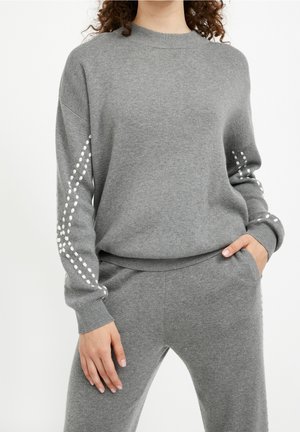 Femme portant un pull gris en maille et un pantalon assorti avec des motifs à pois blancs le long des manches, debout avec une main dans la poche.