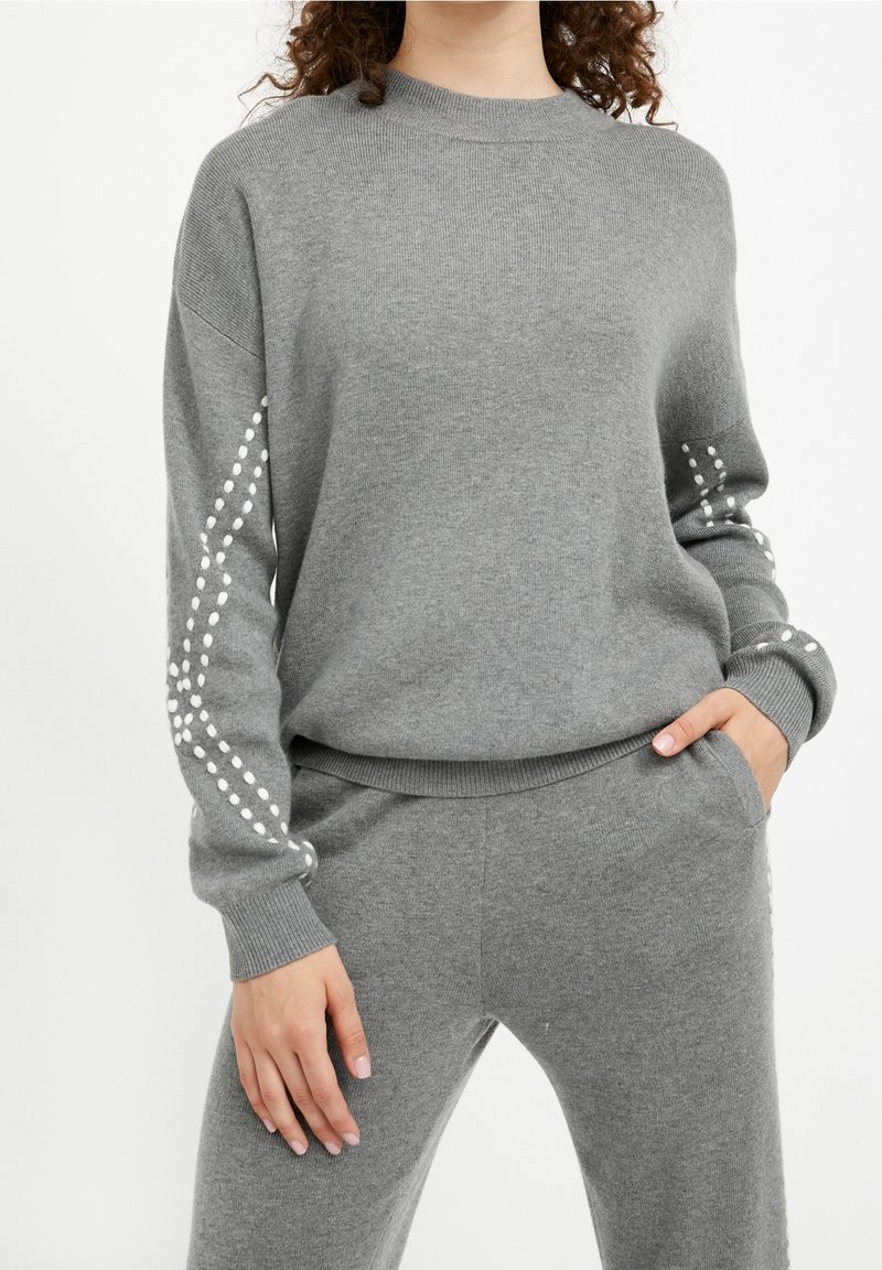 Femme portant un pull gris en maille et un pantalon assorti avec des motifs à pois blancs le long des manches, debout avec une main dans la poche.