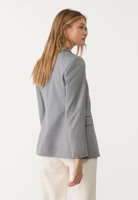 Šedý blazer s texturovanou tkaninou, uvolněný střih a boční kapsy. Dlouhé rukávy a přizpůsobený design doplňují světlé kalhoty.