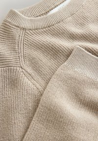 Beige stickad tröja med räfflad textur, med rundad halsringning och kontrasterande släta detaljer vid kragen och ärmsluten.