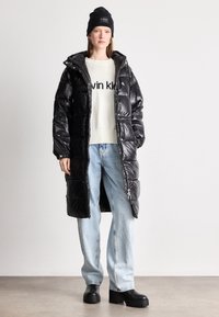 Melnā polsterētā garā mētela ar spīdīgu apdari, ar rāvējslēdzēja priekšpusi un kapuci, uzvilkts virs krēmsvārka ar melnu tekstu un gaiši ziliem džinsiem. Melnas apavus.