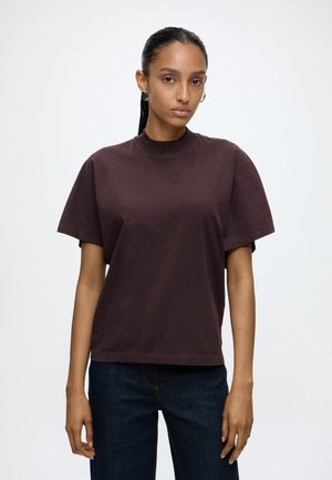 Kobieta z włosami związanymi do tyłu nosi luźny, ciemnobrązowy T-shirt, niebieskie dżinsy i kolczyki-koła, stojąc na tle jasnego, gładkiego tła.