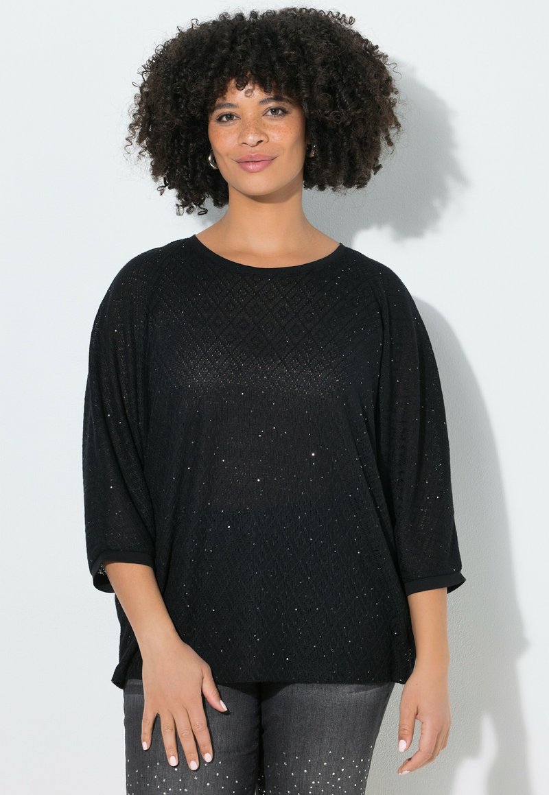 Frau mit lockigem Haar, die einen schwarzen, strukturierten Pullover mit dezenten Glitzerdetails und dunkelgraue Hosen mit verstreuten weißen Punkten trägt.
