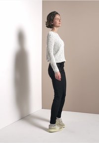 Pull en laine crème, jean noir, baskets grises. Le modèle se tient de côté contre un fond neutre, mettant en valeur la coupe et la texture de la tenue.