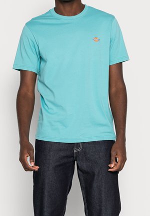Turquoise cotton T-shirt with a crew neck, short sleeves, and a logo accent on the left chest. Paired with dark denim jeans.