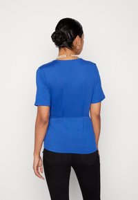 Top azul de mangas curtas com decote redondo, cintura ajustada e tecido suave. Calças pretas e colar acrescentam toques subtis.