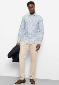 Mann, der ein hellblaues, geblümtes Hemd mit Knöpfen und beige Hosen trägt, hält eine dunkle Jacke und steht vor einer schlichten weißen Wand.