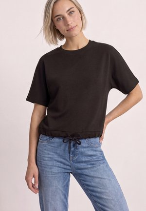 Jeune femme aux cheveux blonds courts portant un haut noir à manches courtes avec un cordon à la taille et un jean bleu clair sur un fond uni.