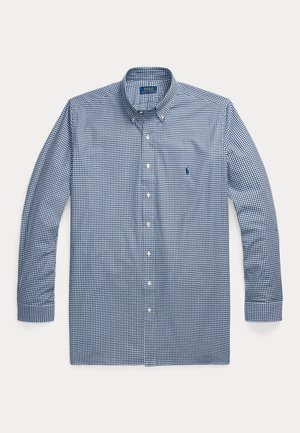 Hombre con camisa azul de cuadros, corbata oscura, vaqueros oscuros, cinturón marrón, zapatos marrones y reloj de pulsera, de pie frente a un fondo beige liso.
