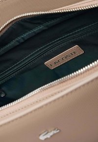 Intérieur d'un sac Lacoste avec une doublure noire lisse, une poche zippée beige et une étiquette en cuir beige. Doté d'une fermeture éclair argentée et d'un extérieur texturé.