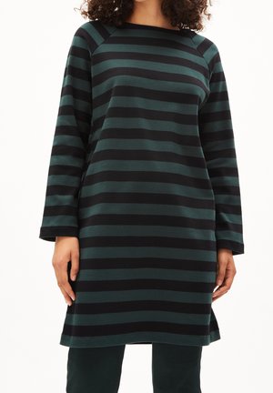 Kobieta ubrana w sukienkę z długim rękawem, sięgającą kolan, w poziome czarne i ciemnozielone paski, stojąca z rękami swobodnie opuszczonymi wzdłuż ciała.