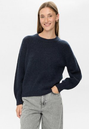 Marinblå stickad tröja med rund halsringning, avslappnad passform och nedhasade axelsömmar, parat med ljusgrå denimjeans.