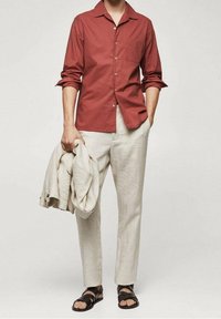 Rood overhemd met knopen, beige linnen broek, een licht jack vasthoudend, zwarte sandalen. Het overhemd heeft een kraag, lange mouwen en een zak.