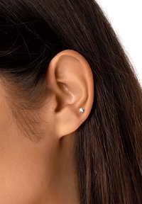 Orecchino a chiodo in argento con una pietra di zirconia cubica trasparente, incastonata in un semplice bezel rotondo. Indossato in un lobo d'orecchio perforato con i capelli scuri parzialmente visibili.