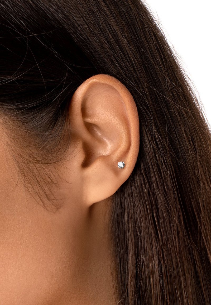 Orecchino a chiodo in argento con una pietra di zirconia cubica trasparente, incastonata in un semplice bezel rotondo. Indossato in un lobo d'orecchio perforato con i capelli scuri parzialmente visibili.