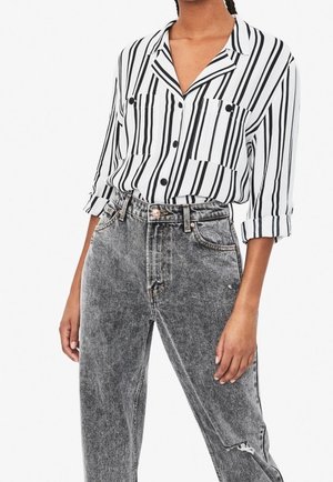 Femme portant une chemise à rayures verticales noires et blanches, boutonnée, aux manches retroussées, et un jean noir délavé taille haute avec une petite déchirure.