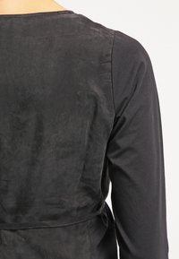 Vue arrière haute d'une personne portant un gilet en daim noir sur une chemise à manches longues noire, sur un fond uni.