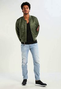 Jaqueta bomber verde-oliva com um bolso com fecho, t-shirt preta, jeans claros com desgaste e sapatilhas pretas. Design de cor sólida.
