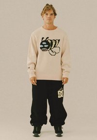 Man in een beige sweater met grote grafische bij en zwarte broek met vlamlogo, staande tegen een effen beige achtergrond.