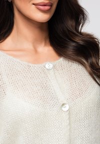Cardigan léger, tricoté de couleur crème avec une texture transparente, doté de deux gros boutons ronds et translucides.
