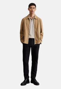 Beige suède jas over een lichtgrijze polo, gecombineerd met zwarte broek en zwarte schoenen. Model staat met handen in de zakken tegen een neutrale achtergrond.