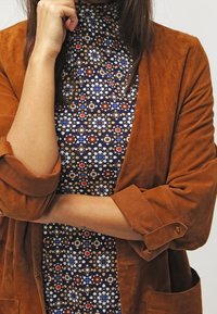 Hnědá semišová bunda s vyhrnutými rukávy přes vzorované šaty s modrými, červenými a bílými geometrickými vzory na tmavě modrém pozadí.