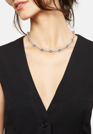 Zilveren ketting met aan elkaar geschakelde ovale vormen, gladde textuur, gedragen om de nek. Gecombineerd met een diep V-hals zwart kledingstuk.