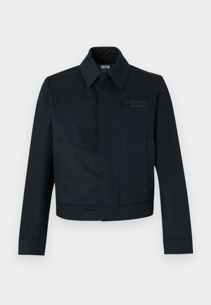 Juoda Versace Jeans Couture striukė su ilgomis rankovėmis, smailiais atvartais, slepiama užsegimu priekyje ir krūtinės emblema dešinėje pusėje.