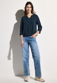 Marineblauwe blouse met een V-hals en trekkoordhals, gecombineerd met lichtblauwe wijdere jeans en beige sneakers. Zachte stof, relaxed pasvorm.