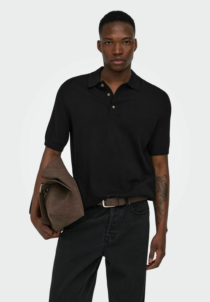 Homme aux cheveux courts, portant un polo noir et un jean noir, tient un sac en cuir marron, montrant un tatouage sur l'avant-bras gauche.