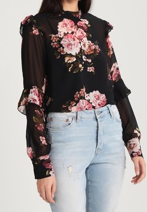 Zwarte doorschijnende blouse met roze bloemenprint en ruches aan de mouwen, gecombineerd met lichtblauwe gescheurde spijkerbroek bij een staand persoon.