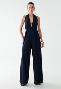 Tamsiai mėlynas kombinezonas su halterio kaklu, giliu V formos iškirpte priekyje, plačiomis kelnėmis ir minkštu, tekstūruotu audiniu. Yra šoninės plyšiai ir minimalūs metaliniai elementai.