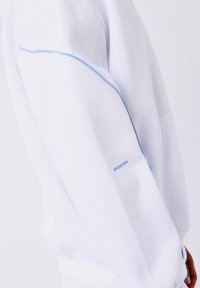 Witte sweatshirt mouw met lichtblauwe biezen langs de arm en kleine blauwe "PXP" tekst nabij de pols.