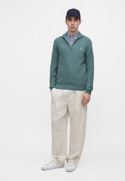 Jeune homme portant une casquette bleu foncé, un pull vert à fermeture éclair partielle sur une chemise à carreaux, un pantalon beige ample et des baskets blanches, debout sur un fond blanc.
