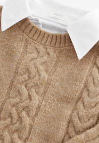 Beige kötött pulóver, amely kábelmintával és kerek nyakkivágással rendelkezik, egy fehér galléros ing fölé rétegezve. Textúrált gyapjú anyag.