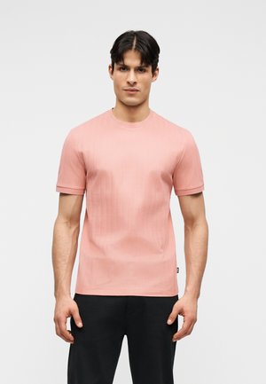 Giovane uomo di fronte che indossa una maglietta rosa a maniche corte aderente a coste e pantaloni neri su uno sfondo chiaro e uniforme.